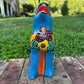 A hand-painted talavera ceramic dog figurine in a sitting position, featuring a vibrant blue base with colorful geometric patterns in red, yellow, orange, and white. The figurine is photographed outdoors on a brick surface with a blurred green lawn and trees in the background front view