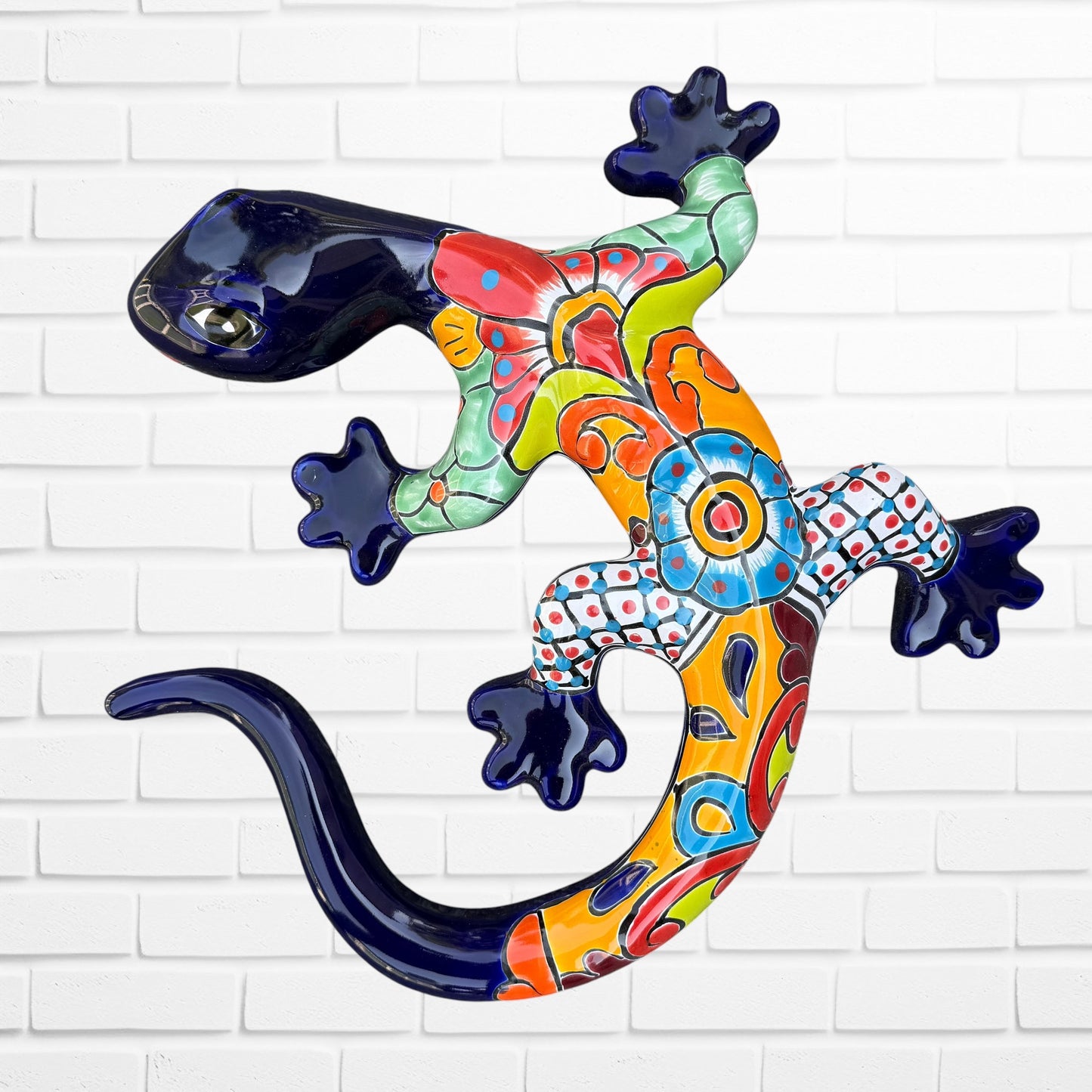 A colorful Mexican Talavera wall sculpture of a lizard, featuring a vibrant multicolor pattern with dark blue, orange, red, green, and yellow, mounted on a white brick wall.