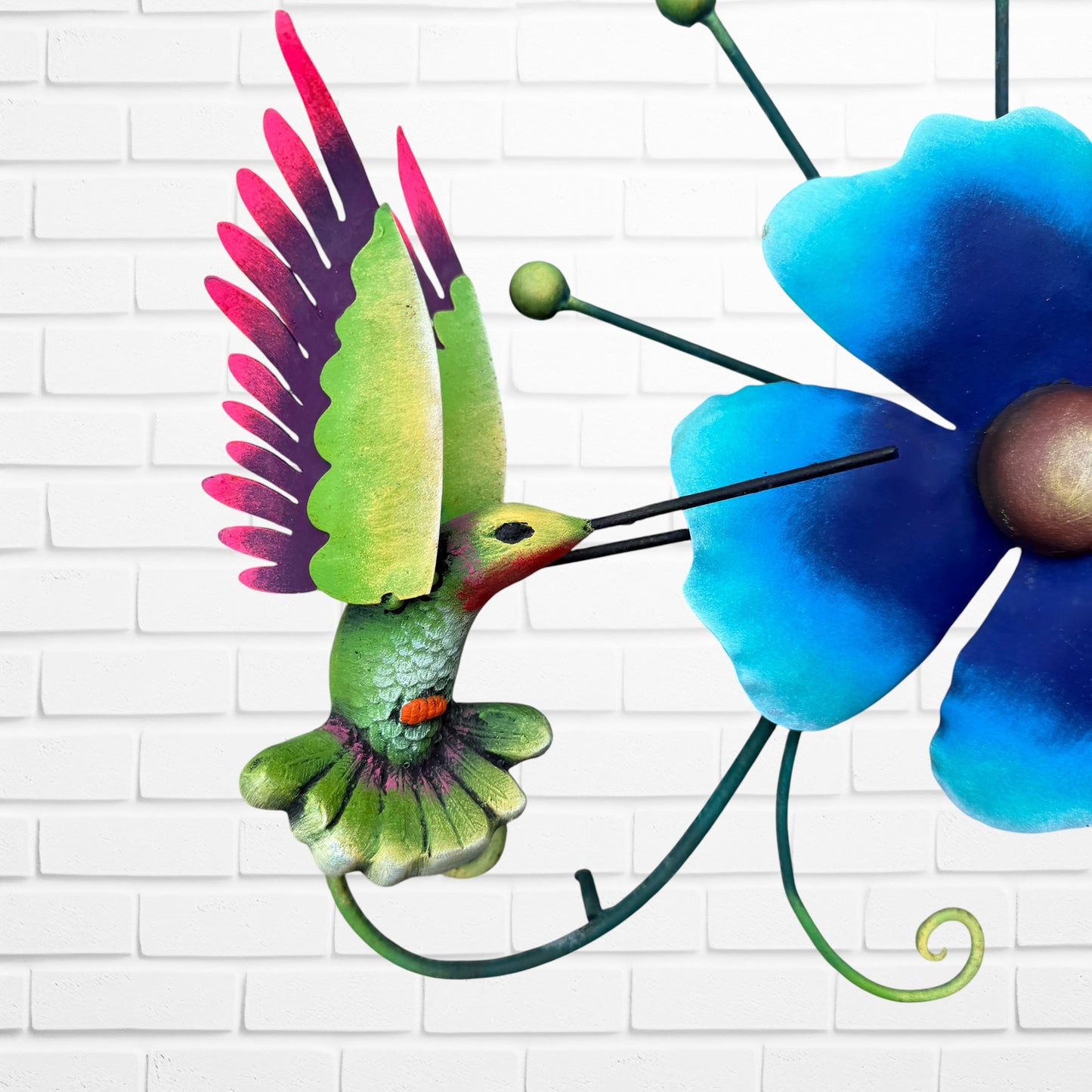 Decorative metal wall art with three blue flowers and a colorful hummingbird on a white brick background bird close up