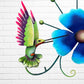Decorative metal wall art with three blue flowers and a colorful hummingbird on a white brick background bird close up