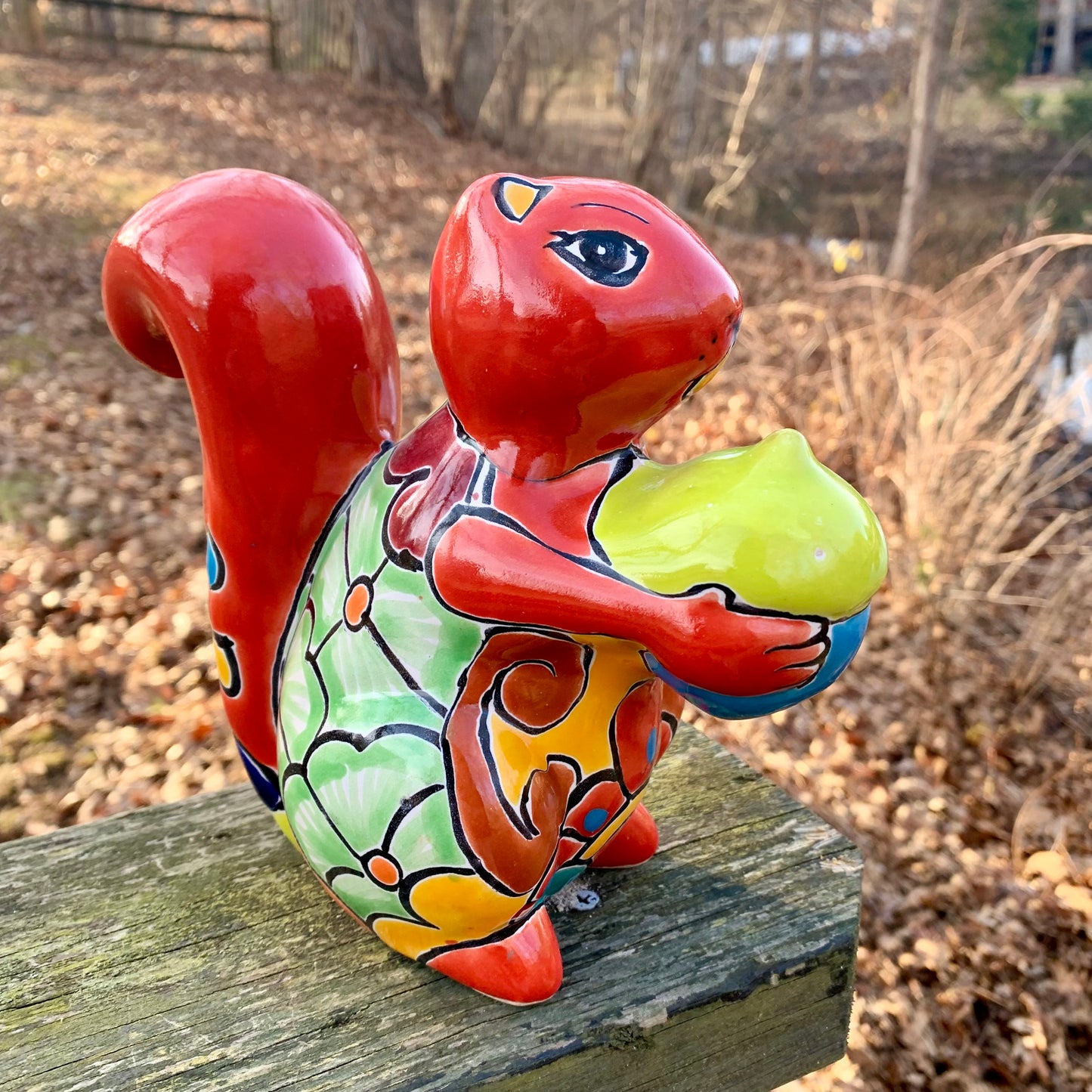 Colorful ceramic squirrel figurine holding a nut on a wooden surface with a natural background in red.