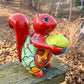 Colorful ceramic squirrel figurine holding a nut on a wooden surface with a natural background in red.