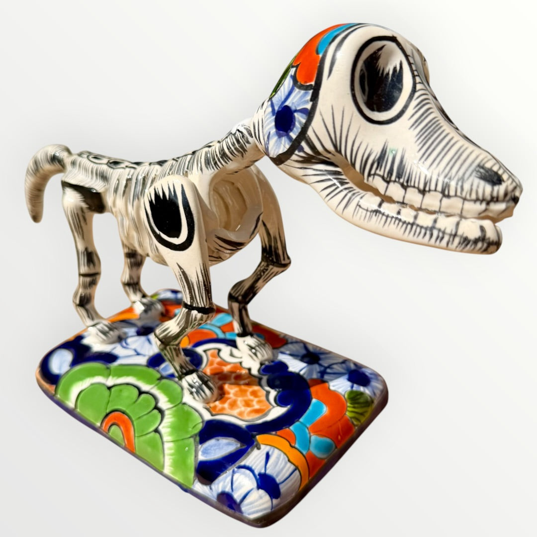 A ceramic Mexican-style skeleton dog figurine standing on a colorful floral base against a plain white background.