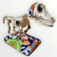 A ceramic Mexican-style skeleton dog figurine standing on a colorful floral base against a plain white background.