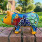 A colorful ceramic pig planter with a yellow-orange face and body, decorated with blue, green, red, and white floral patterns, sitting on a brick surface with a blurred outdoor garden background. Photo of right side view.