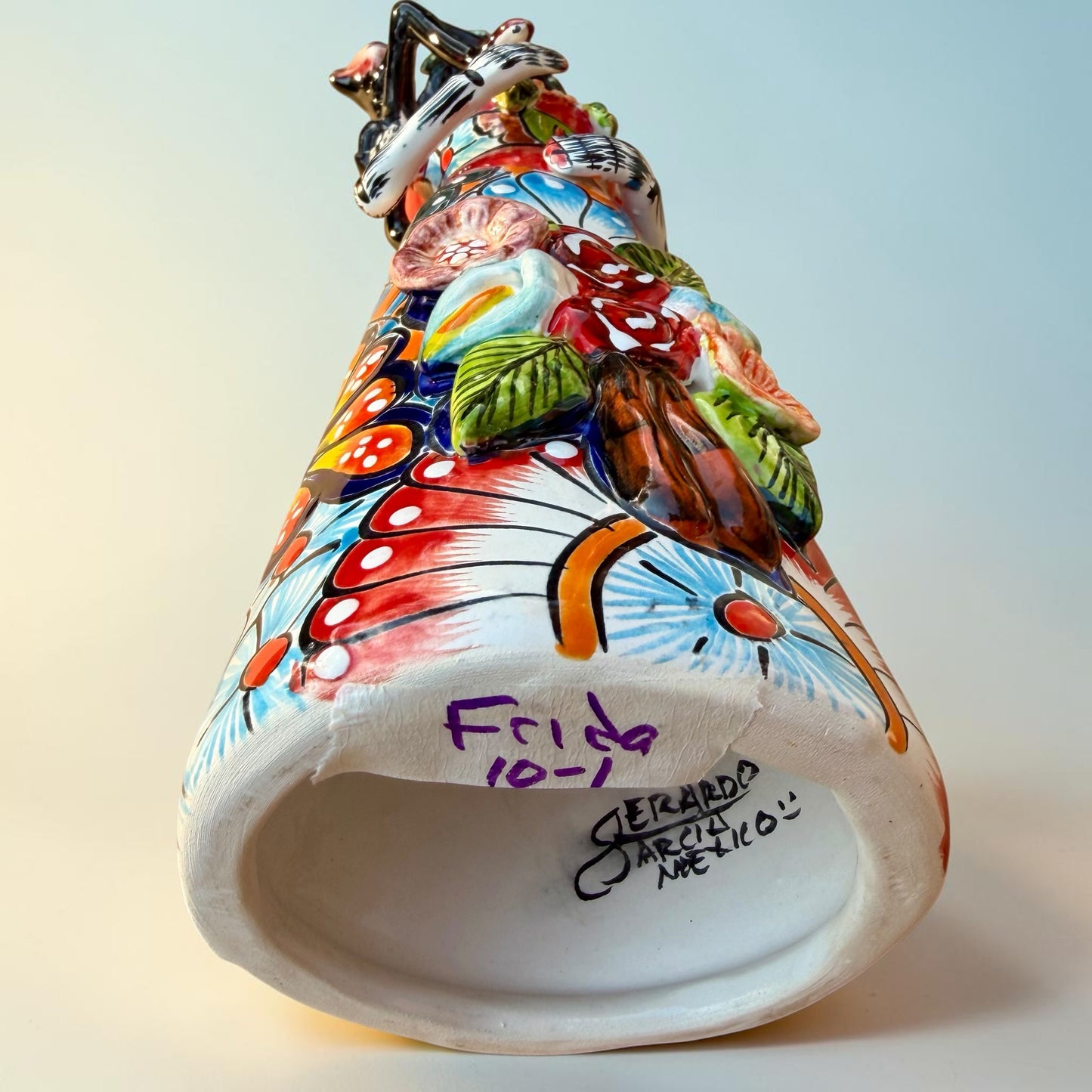 A Talavera Day of the Dead (Día de los Muertos) Frida figurine  with a monkey on her shoulder and a red parrot on her other shoulder, set against a light blue gradient background. Bottom view showing artist signature.