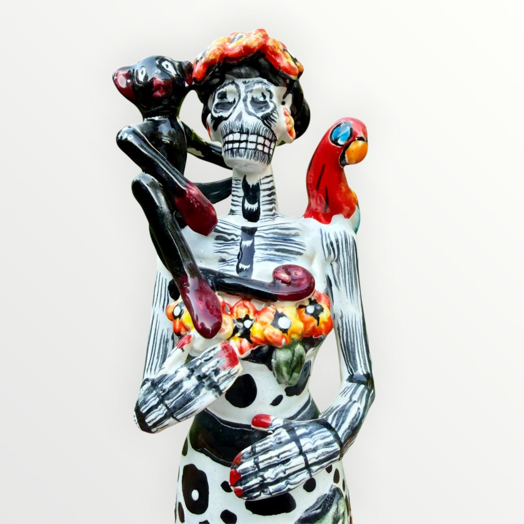 A Day of the Dead Catrina of a Frida skeleton doll holding a black cat, with a red parrot on her shoulder and colorful flowers around the top of her dress.