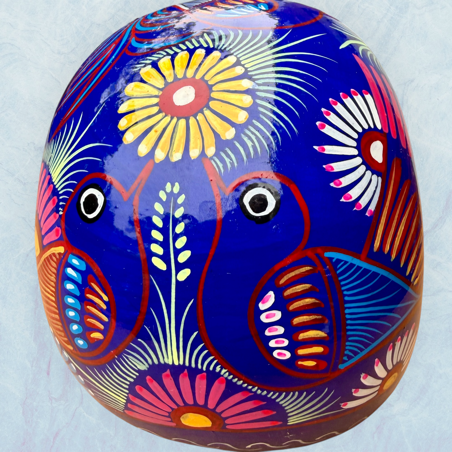 A glossy blue Day of the Dead sugar skull XGrande decorated with vibrant pink, blue, white, and orange floral and geometric patterns, shown against a light blue textured background, back view.