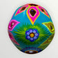 A decorative sugar skull painted in bright turquoise blue with colorful floral and geometric patterns in pink, yellow, green, purple, and white, shown against a plain light background back view
