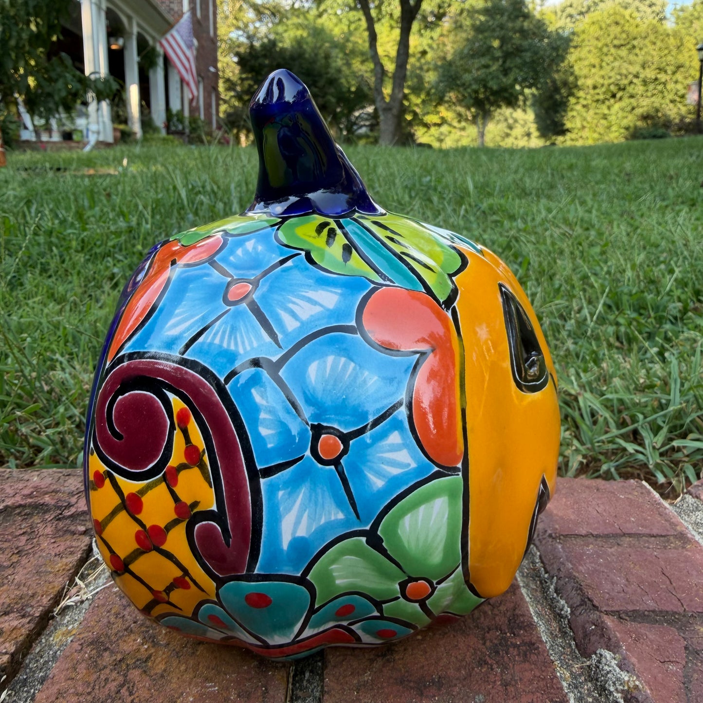 A Talavera jack-o'-lantern pumpkin decoration with a carved face, sitting on a brick surface in an outdoor yard with grass and trees in the background. Left side view of pumpkin.