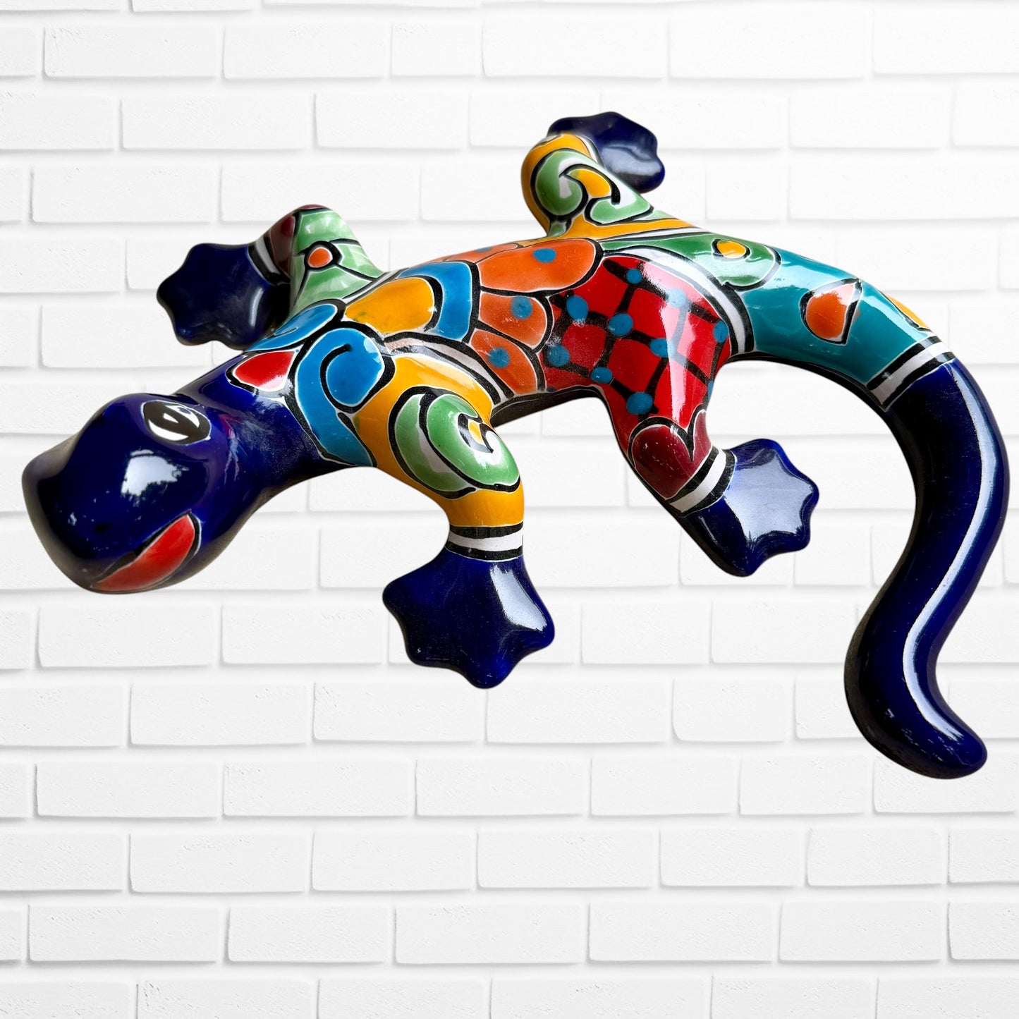 A colorful ceramic wall sculpture of a stylized lizard, decorated with vibrant hand-painted patterns in blue, orange, green, yellow, and red, mounted on a white brick wall front view