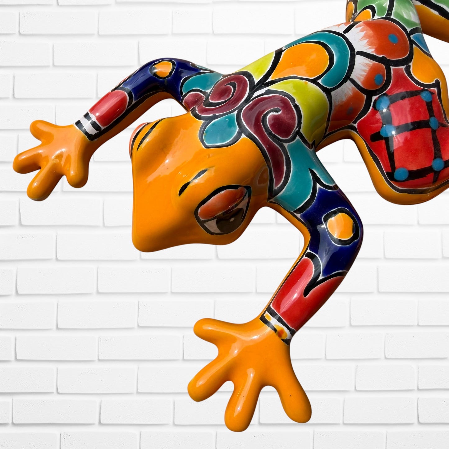 A colorful hand-painted ceramic frog sculpture in a dynamic crawling pose, set against a white painted brick wall background.