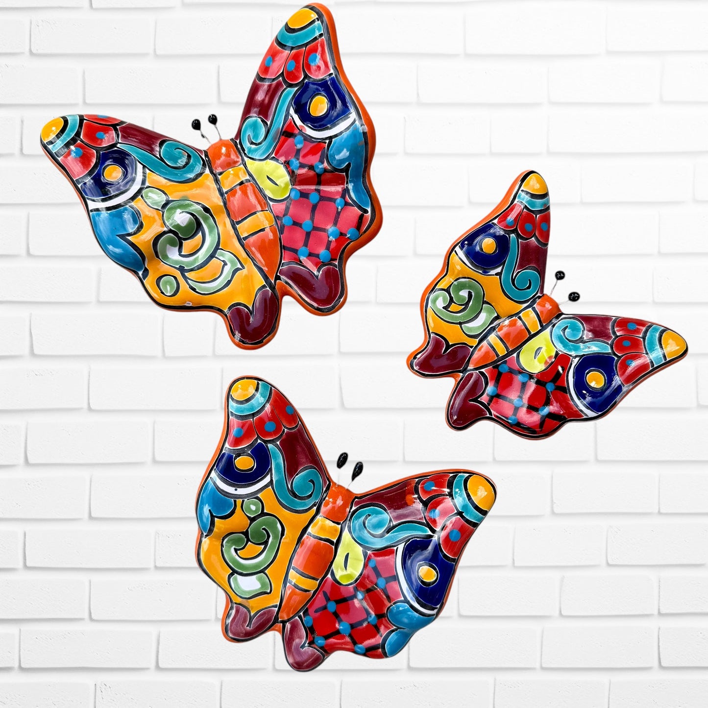 Three colorful talavera butterfly wall art pieces with orange and traditional multicolor patterns, displayed against a white brick wall. 