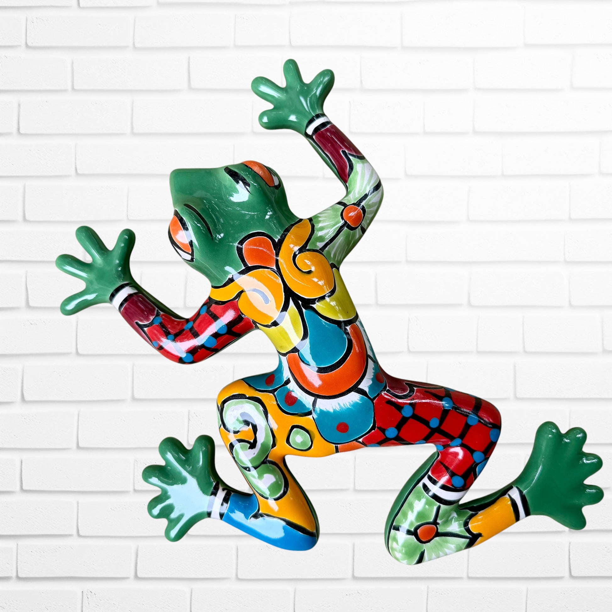 A colorful hand-painted ceramic frog wall sculpture in a dynamic jumping pose, decorated with vibrant green, red, orange, yellow, and blue patterns, set against a white brick wall background.