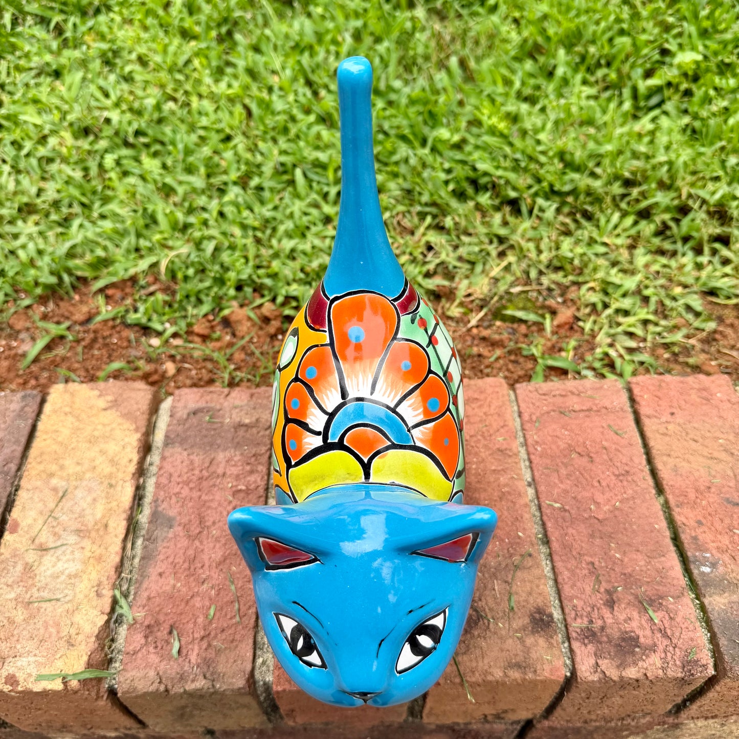 A glossy talavera ceramic cat figurine decorated with vibrant hand-painted patterns in turquoise, yellow, green and red, standing on a brick path with a green lawn and a house in the blurred background top kitty view.