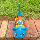 A glossy talavera ceramic cat figurine decorated with vibrant hand-painted patterns in turquoise, yellow, green and red, standing on a brick path with a green lawn and a house in the blurred background top kitty view.