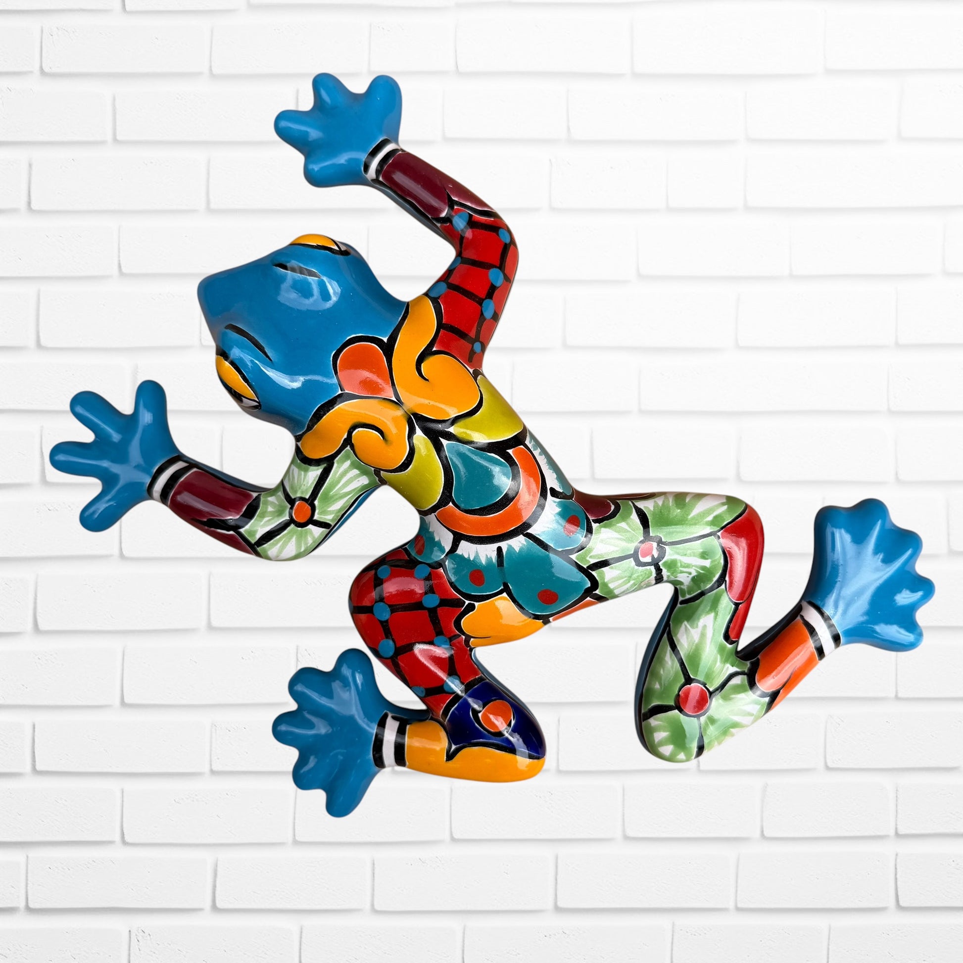 A colorful hand-painted talavera blue frog wall sculpture in a dynamic jumping pose, mounted on a white painted brick wall.