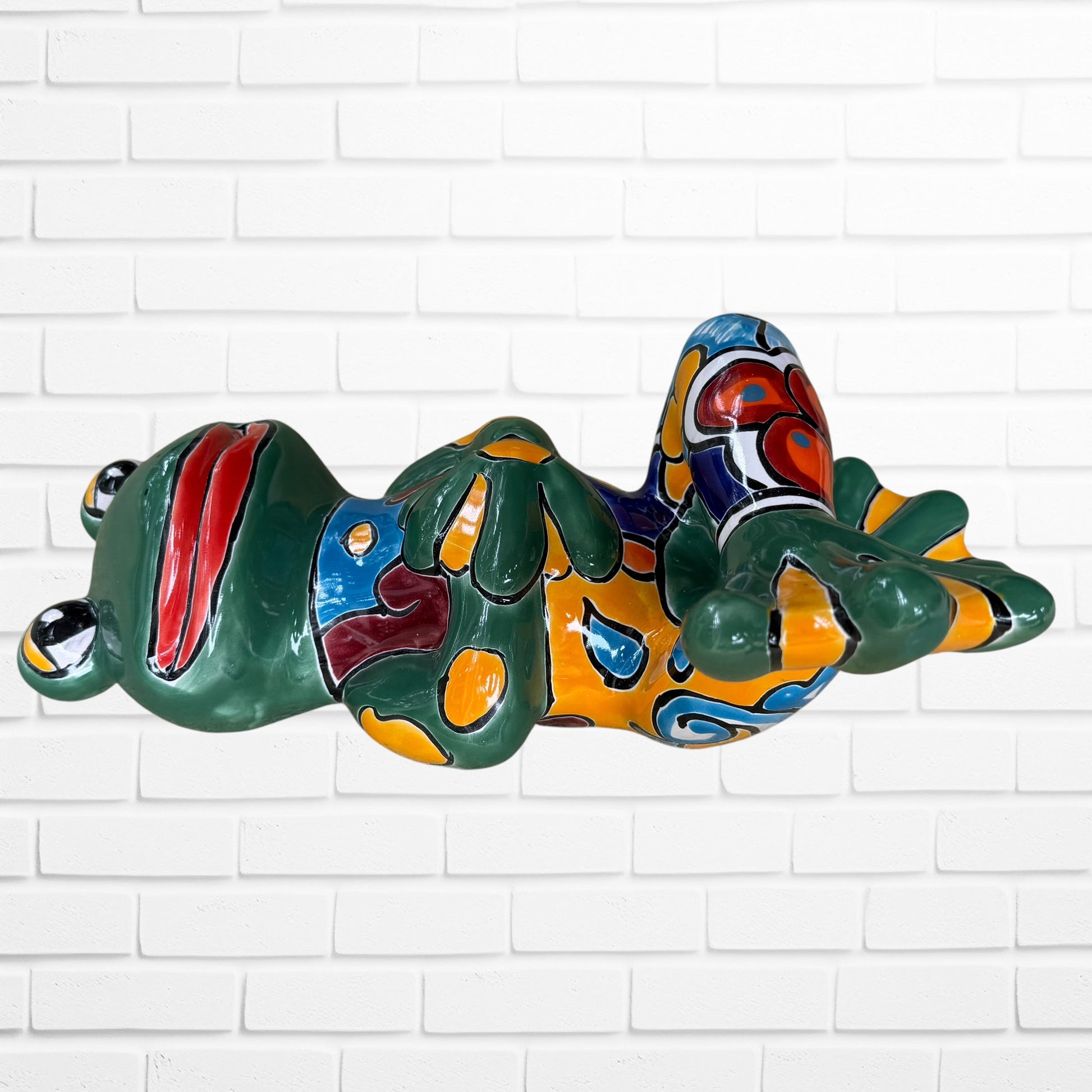 A colorful ceramic frog wall sculpture sitting cross-legged against a white painted brick wall. The frog features a glossy green body with yellow, blue, red, and Right side view