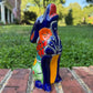 A colorful hand-painted Mexican Talavera Ceramic dog figurine sitting on a brick wall with a green grassy lawn and a house in the background.