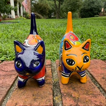 Two colorful talavera ceramic cat figurines standing on a brick surface outdoors with grass and trees blurred in the background. One cat is dark blue with orange, yellow, and red patterns, and the other is orange with blue, red, yellow, and white patterns. This is the front view.