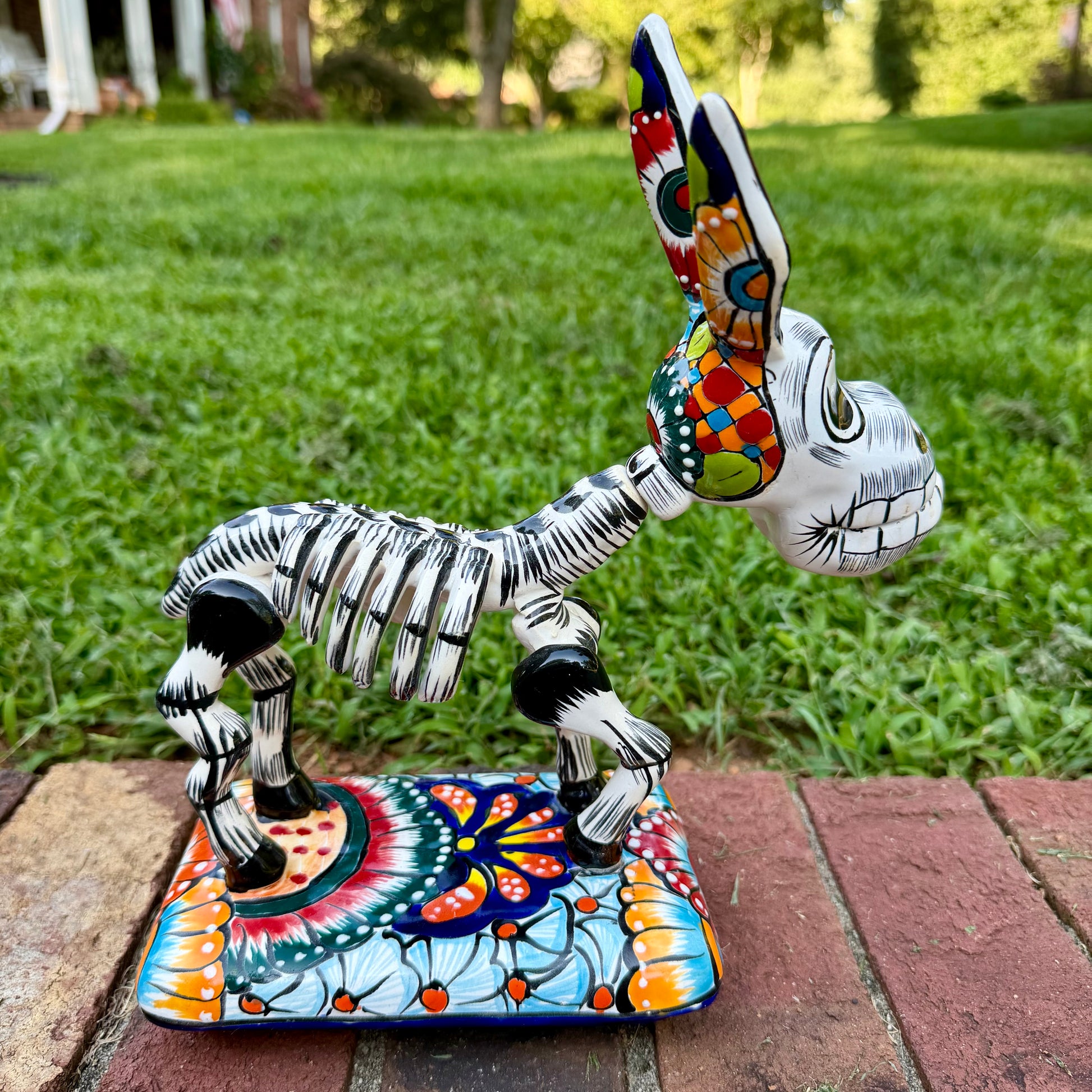 A colorful Catrina donkey figurine standing on a decorated base, photographed outdoors on a brick surface with a grassy yard and house in the background, left side view of donkey. 