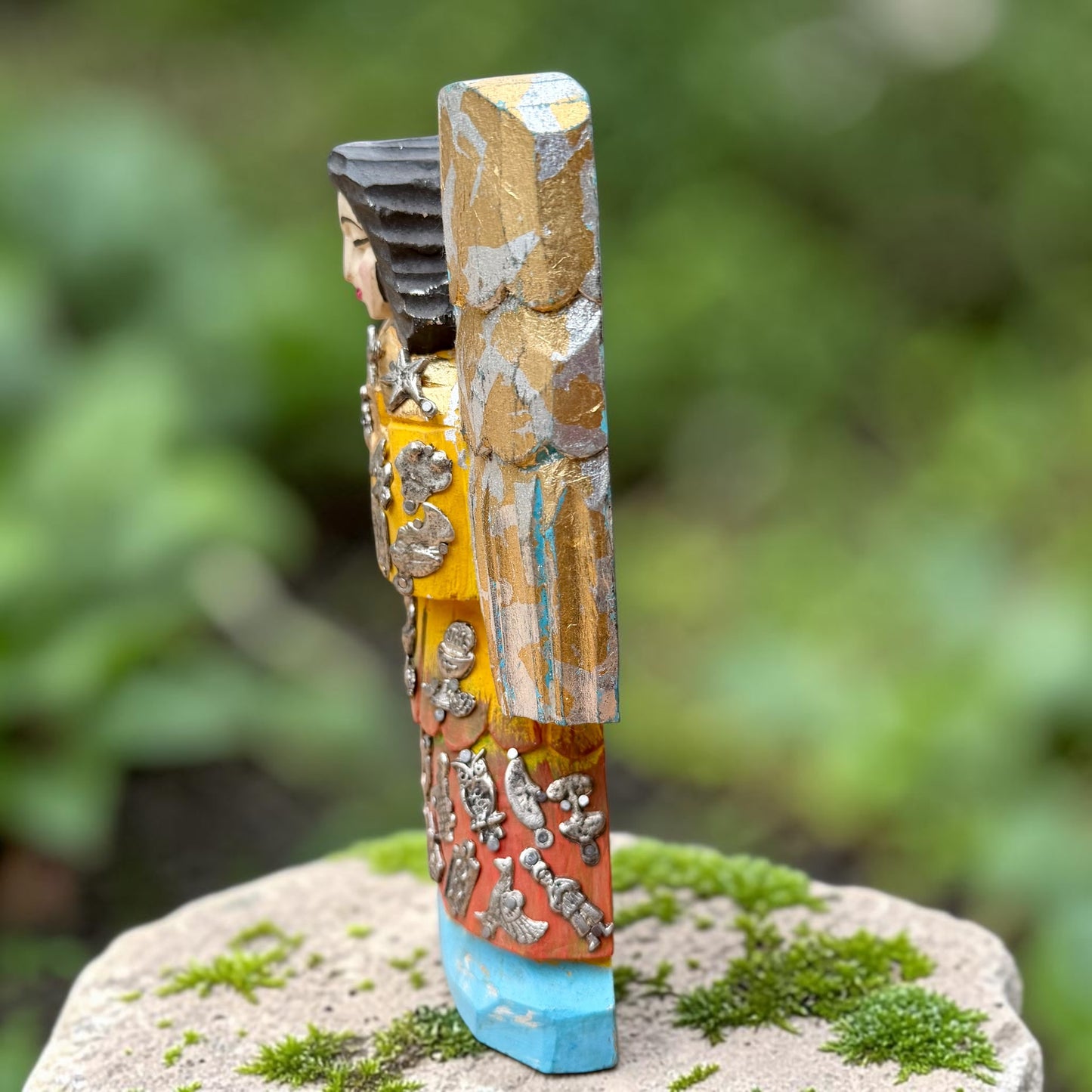 A handcrafted wooden Milagros angel figurine with large wings, decorated with metallic silver charms and painted in yellow, and orange, standing on a mossy stone with a blurred green garden background. Right Side view of angel.