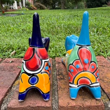 Two colorful hand-painted ceramic cats sitting on a brick ledge with a green lawn and trees in the background. One is cobalt  blue and the other is turquoise, both have traditional multi-color  talavera designs, back view.