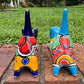 Two colorful hand-painted ceramic cats sitting on a brick ledge with a green lawn and trees in the background. One is cobalt  blue and the other is turquoise, both have traditional multi-color  talavera designs, back view.