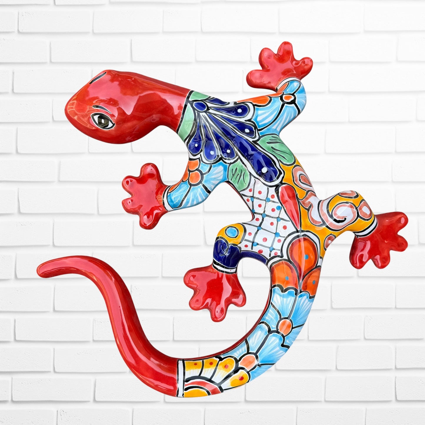 A colorful Talavera ceramic wall salamander lizard art piece with a glossy finish, featuring a red head and tail with a patterned body, displayed against a white brick wall background.