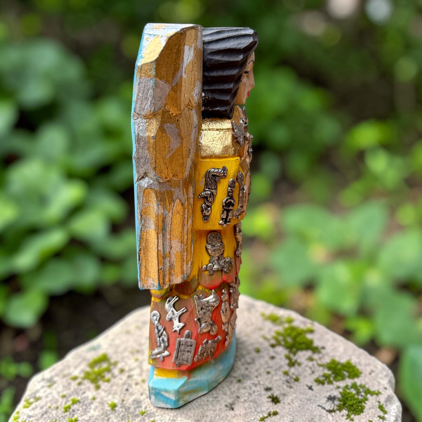 A handcrafted wooden Milagros angel figurine with large wings, decorated with metallic silver charms and painted in yellow, and orange, standing on a mossy stone with a blurred green garden background. Left side view of angel.