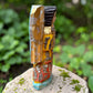 A handcrafted wooden Milagros angel figurine with large wings, decorated with metallic silver charms and painted in yellow, and orange, standing on a mossy stone with a blurred green garden background. Left side view of angel.