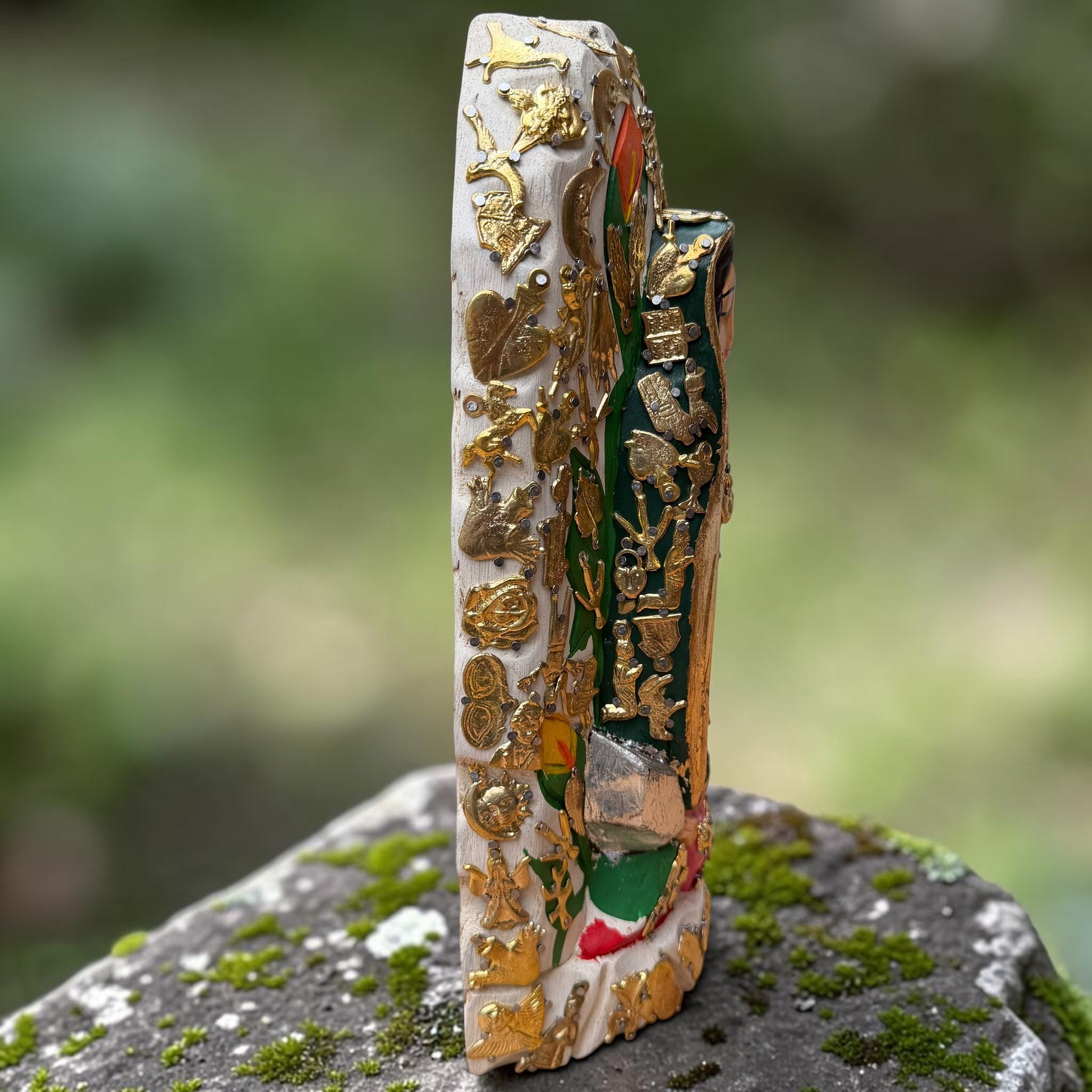 A Milagros religious figurine of Our Lady of Guadalupe with a child, standing on a mossy stone surface with a blurred outdoor garden background. She has a white halo and Lillies adorning the halo. left side view.