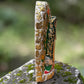 A Milagros religious figurine of Our Lady of Guadalupe with a child, standing on a mossy stone surface with a blurred outdoor garden background. She has a white halo and Lillies adorning the halo. left side view.