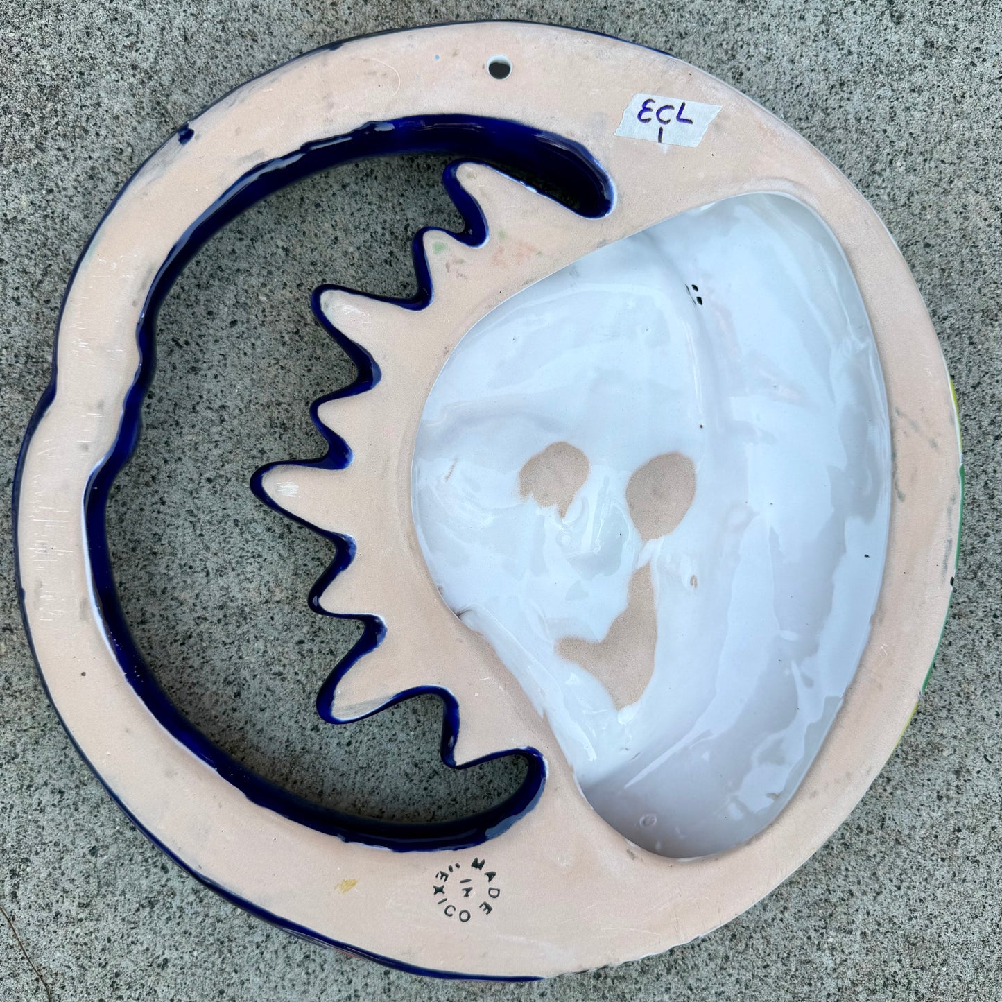 A colorful talavera wall hanging plaque featuring a stylized sun and moon face with decorative floral and star patterns, photographed from above on a gray stone surface, back view Showing hanging hole.