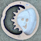 A colorful talavera wall hanging plaque featuring a stylized sun and moon face with decorative floral and star patterns, photographed from above on a gray stone surface, back view Showing hanging hole.
