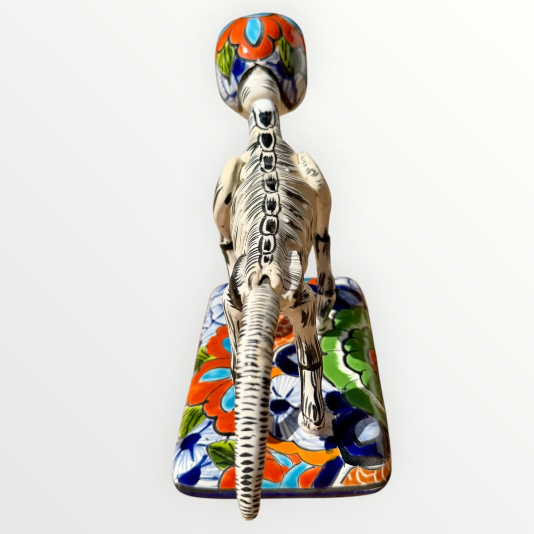 A ceramic Mexican-style skeleton dog figurine standing on a colorful floral base against a plain white background. Back view of dog showing backbone.