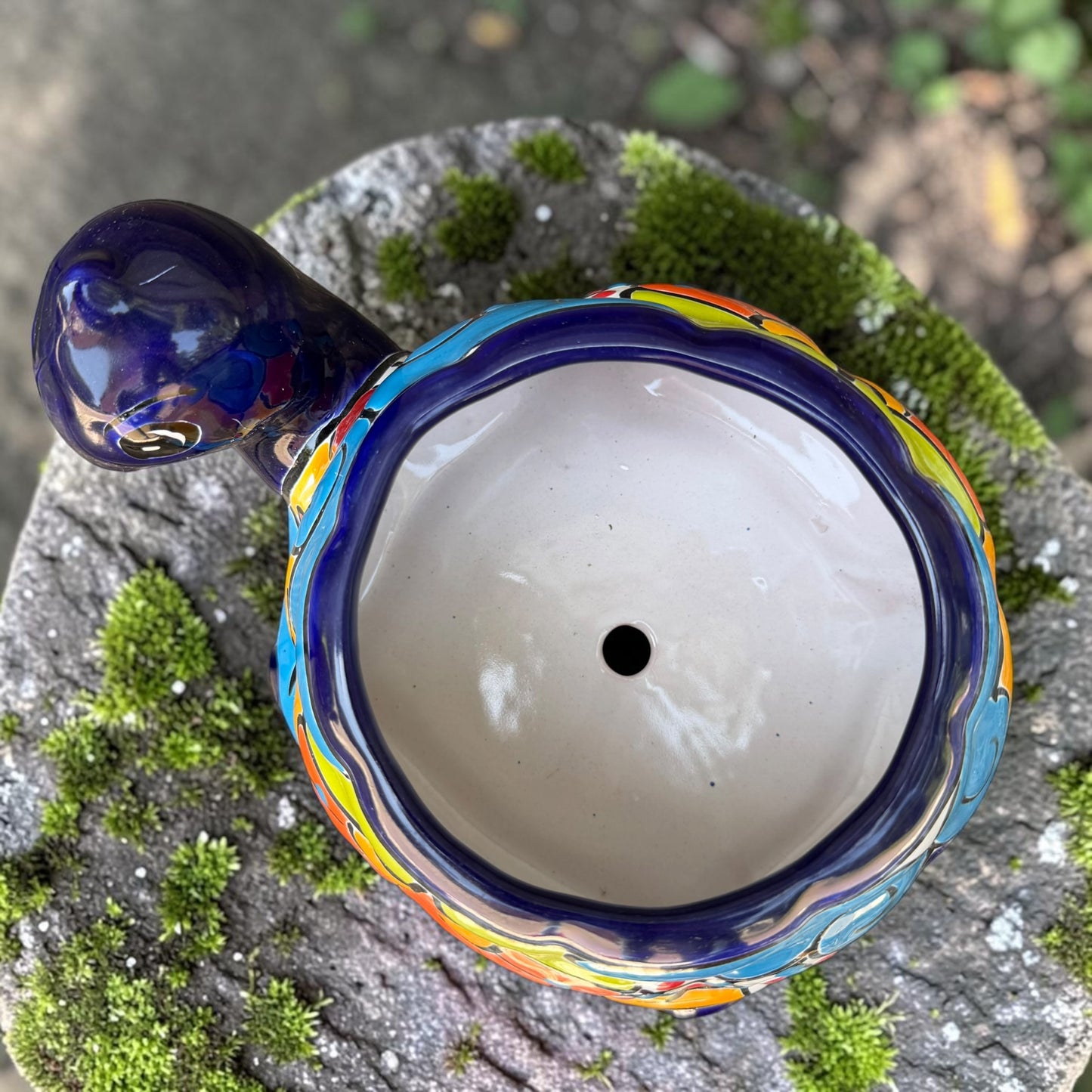 A colorful talavera turtle planter with a hand-painted Mexican-style pattern in orange, blue, yellow, and white, sitting on a mossy stone outdoors. Top view showing opening for plant.