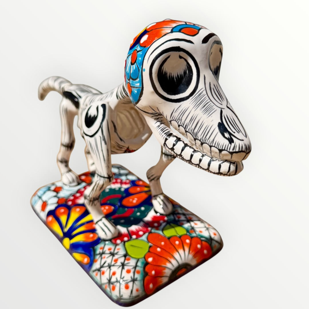 A ceramic Mexican-style skeleton dog figurine standing on a colorful floral base against a plain white background.