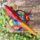 A colorful ceramic rooster figurine standing on a weathered wooden surface outdoors, with a green wooden wall behind it. The rooster is decorated with bright red, yellow, blue, and orange painted details, top view.