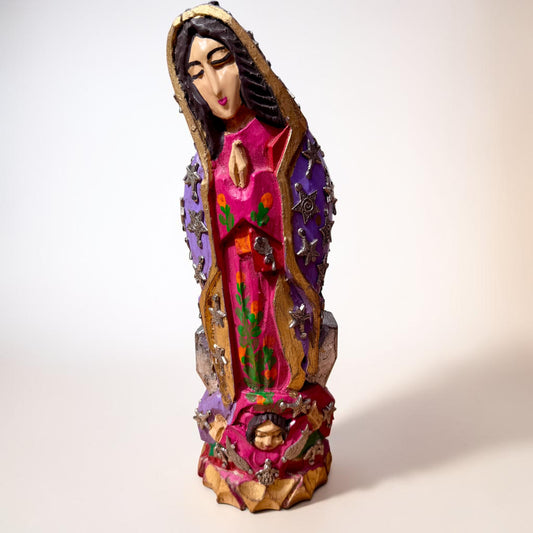A colorful milagros figurine of Our Lady of Guadalupe with a child at her feet, featuring a vibrant pink robe, purple cloak, and gold accents, set against a plain off-white background.