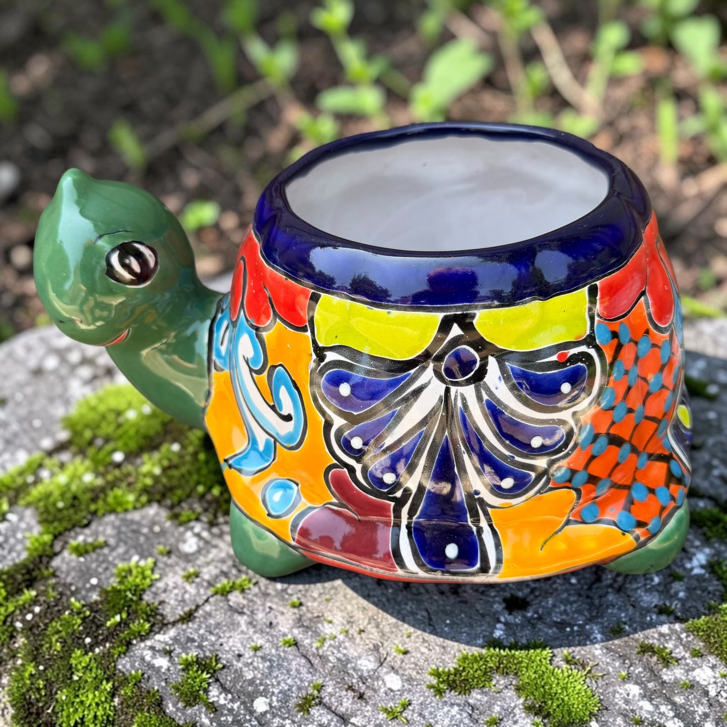 A colorful ceramic turtle-shaped planter with a green head and legs, decorated with vibrant hand-painted patterns in orange, yellow, blue, red, and white, sitting on a mossy stone outdoors.