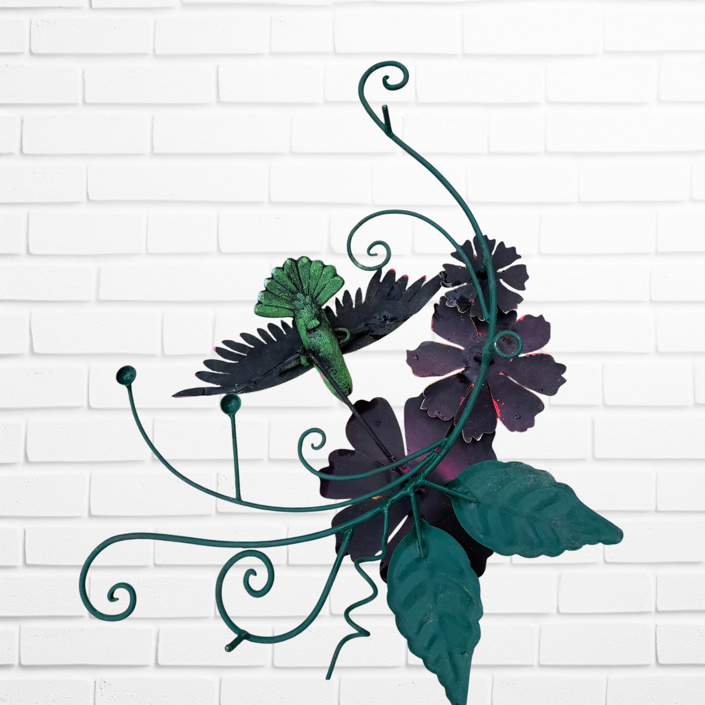 Decorative metal wall art with orange flowers and a hummingbird on a white brick background back