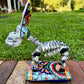 A colorful Catrina donkey figurine standing on a decorated base, photographed outdoors on a brick surface with a grassy yard and house in the background, right side view of body. 