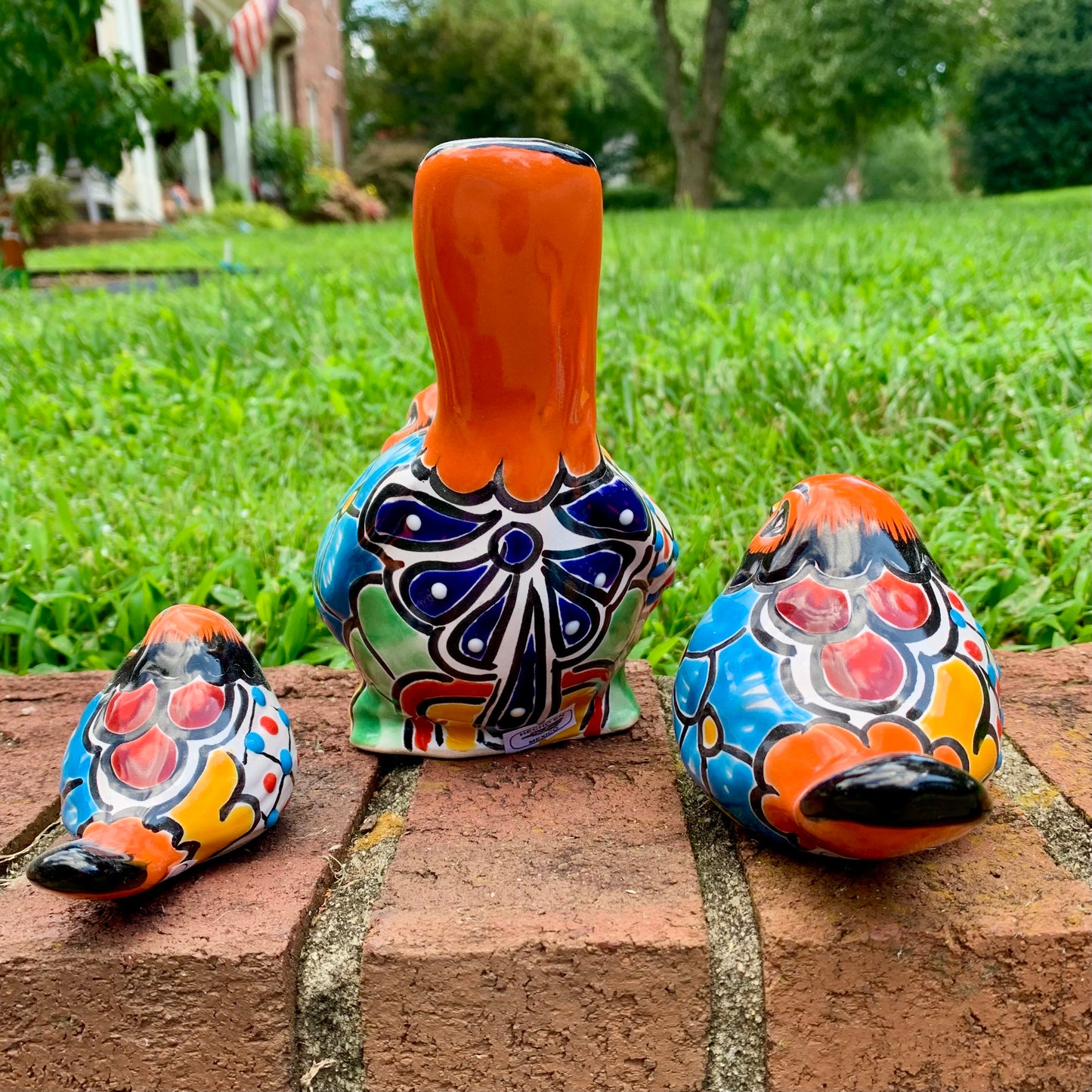 Three hand-painted ceramic bird garden statues in orange, blue, yellow, and black, displayed on a brick ledge in a backyard with a green lawn and trees in the background back view.