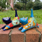 Two colorful hand-painted ceramic cats sitting on a brick ledge with a green lawn and trees in the background. One is cobalt  blue and the other is turquoise, both have traditional multi-color  talavera designs, right side view.