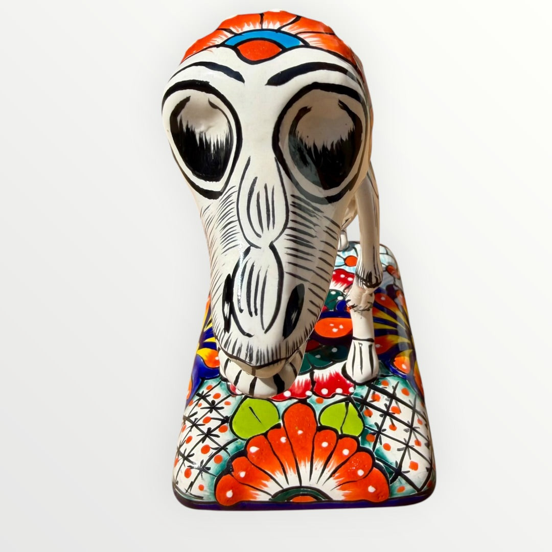 A ceramic Mexican-style skeleton dog figurine standing on a colorful floral base against a plain white background. Close up of front face.