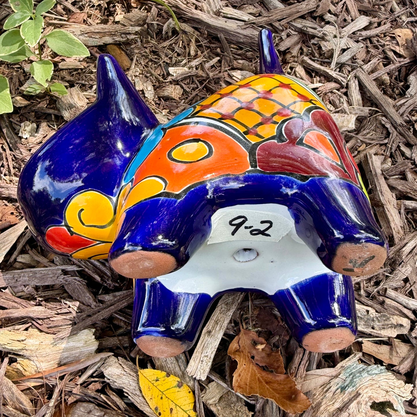 A colorful glazed ceramic cat figurine standing on mulch outdoors, with green plants blurred in the background. The cat is decorated with blue, orange, yellow, and white patterns, bottom view of cat.