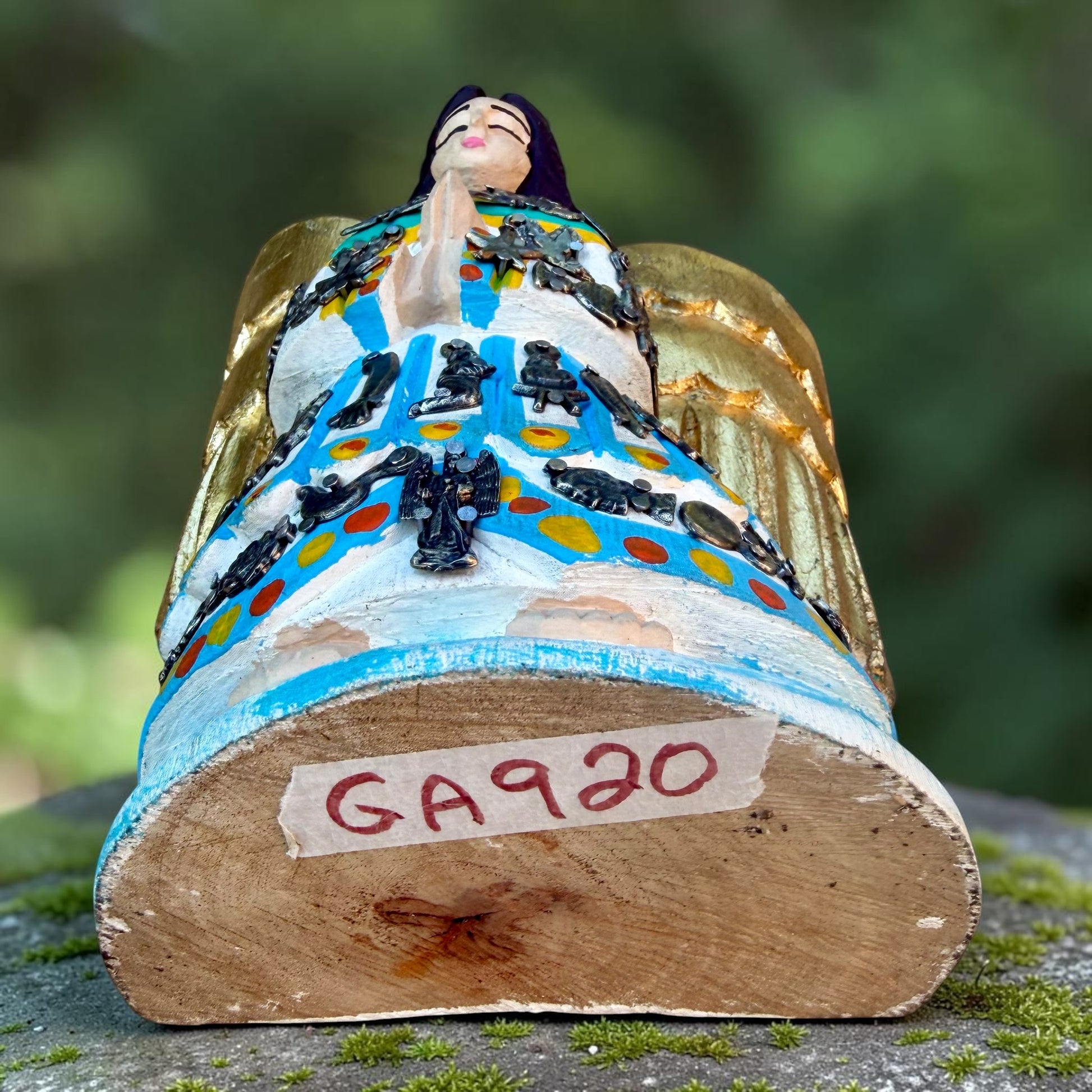 A Milagros guardian angel figurine with gold wings, dark hair, and a blue, white, and gold embroidered dress, posed outdoors on a stone surface with a blurred green background. This is an angled view showing her from the bottom of sculpture up.