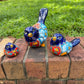 Three hand-painted ceramic bird figurines in different sizes, decorated with a vibrant Mexican Talavera style pattern of blue, red, yellow, and white, displayed on a brick ledge with a green grass background.