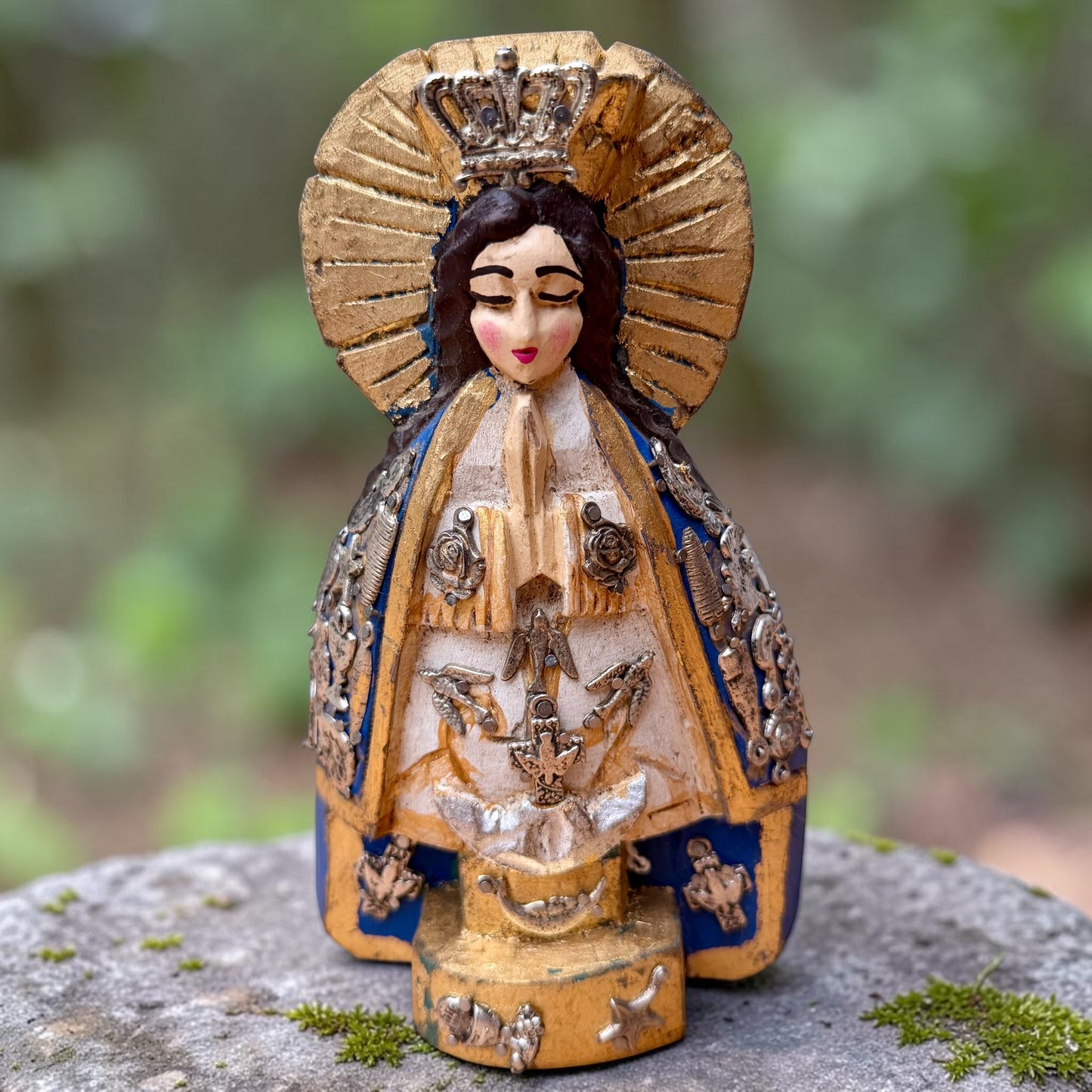 A Milagros figurine of the Virgin Mary Salud with a gold halo and crown, blue and beige robes, and silver-toned decorative details, standing on a stone surface with a blurred green outdoor background.